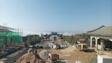 A construction site with traditional Asian architecture in the background, surrounded by scaffolding and building materials. A large piece of construction equipment is visible, and in the distance, a misty landscape can be seen, creating a serene backdrop.