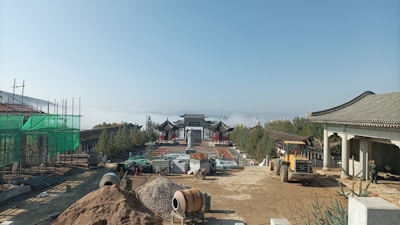 A construction site with traditional Asian architecture in the background, surrounded by scaffolding and building materials. A large piece of construction equipment is visible, and in the distance, a misty landscape can be seen, creating a serene backdrop.