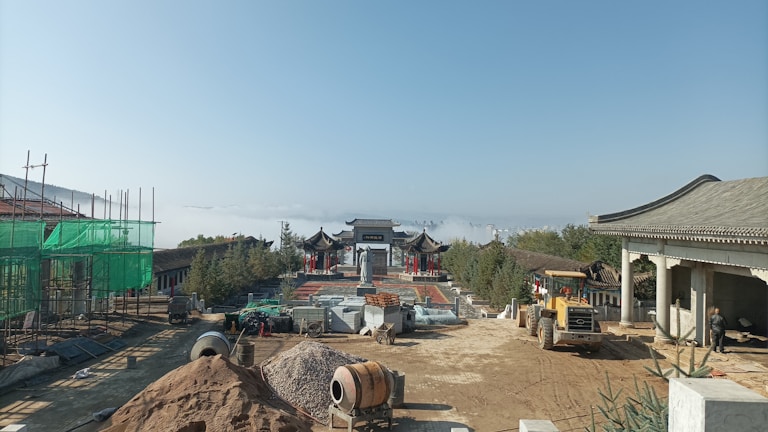 A construction site with traditional Asian architecture in the background, surrounded by scaffolding and building materials. A large piece of construction equipment is visible, and in the distance, a misty landscape can be seen, creating a serene backdrop.