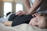 A person is lying face down on a comfortable surface, receiving a massage from another individual's hands. The setting appears calm and serene, with soft lighting enhancing the relaxed atmosphere. The person receiving the massage has closed eyes, indicating a state of relaxation.