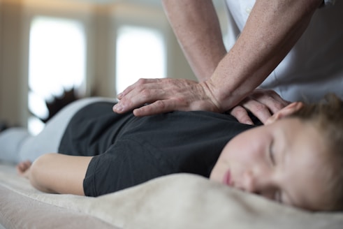 A person is lying face down on a comfortable surface, receiving a massage from another individual's hands. The setting appears calm and serene, with soft lighting enhancing the relaxed atmosphere. The person receiving the massage has closed eyes, indicating a state of relaxation.