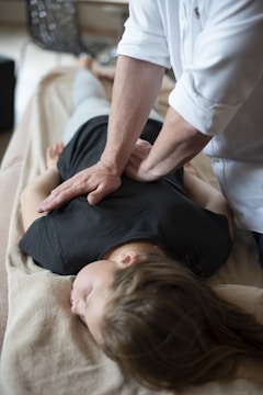 A person lies face down on a massage table while another person applies pressure on their back with both hands. The setting appears calm and the clothes suggest a casual, relaxed environment, possibly in a spa or therapy center.
