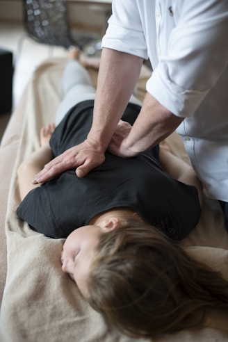 A person lies face down on a massage table while another person applies pressure on their back with both hands. The setting appears calm and the clothes suggest a casual, relaxed environment, possibly in a spa or therapy center.