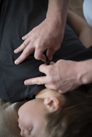Hands appear to be performing a massage technique on a person's back, with fingers pressing into the back near the shoulders. The person receiving the massage is laying on a soft surface with their face turned to the side.
