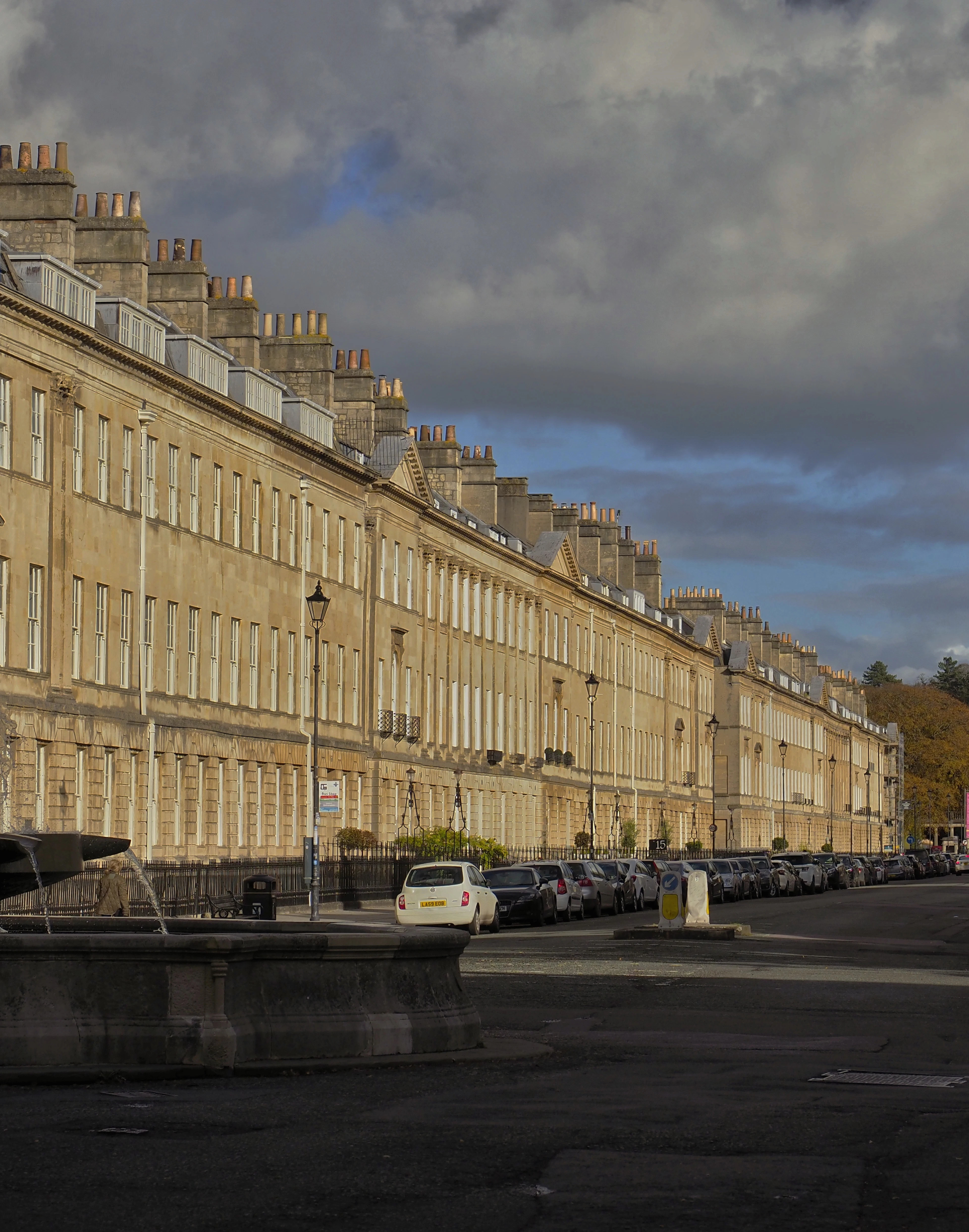 The long Georgian terrace of Great Pulteney Street in Bath under low autumn sun, chimney stacks marching away to the horizon above a stone fountain basin in the foreground.