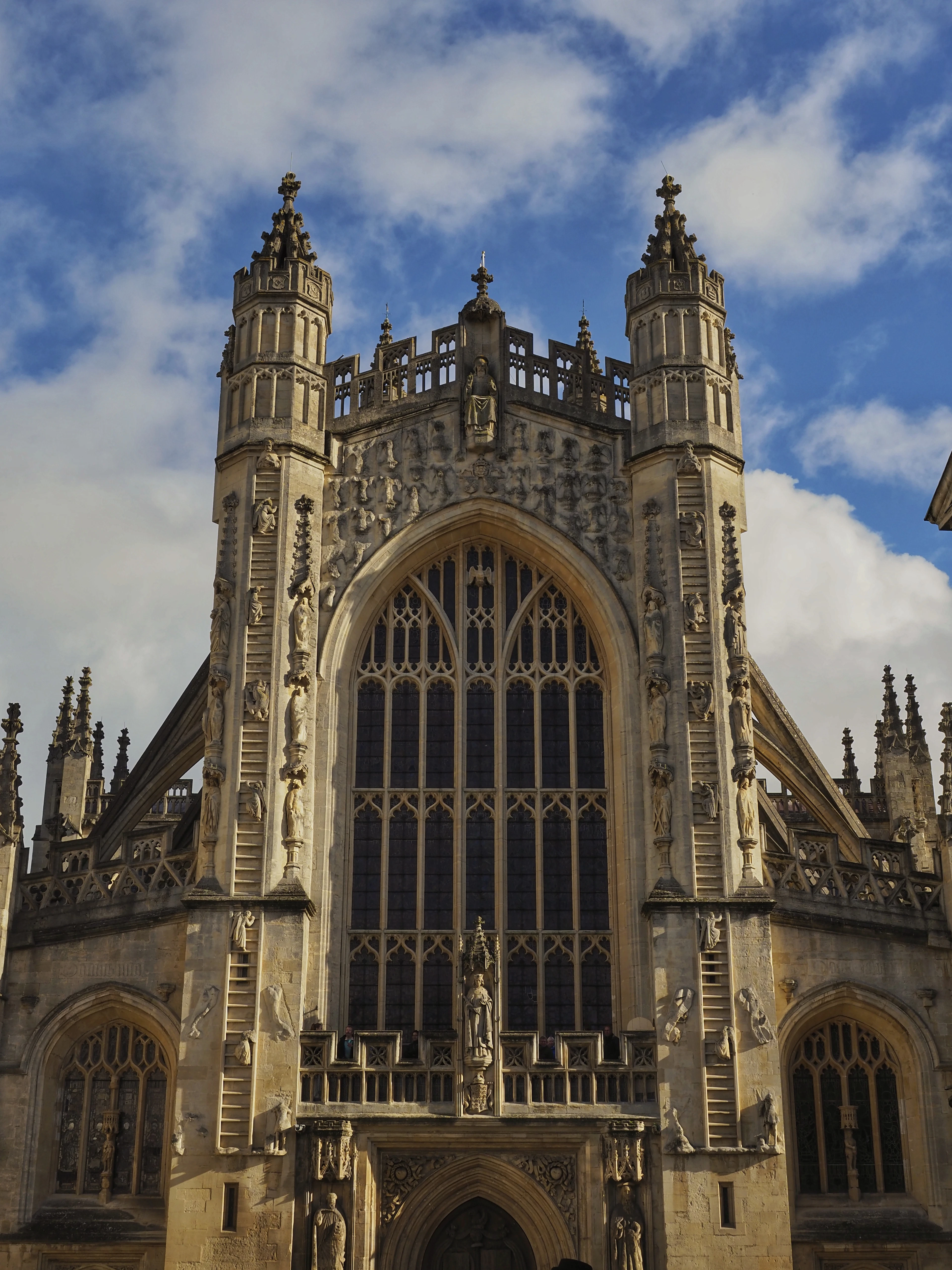 The west front of Bath Abbey seen from below in low sun, its twin turrets carved with the famous stone ladders of climbing angels flanking a vast perpendicular window of honey-coloured limestone.