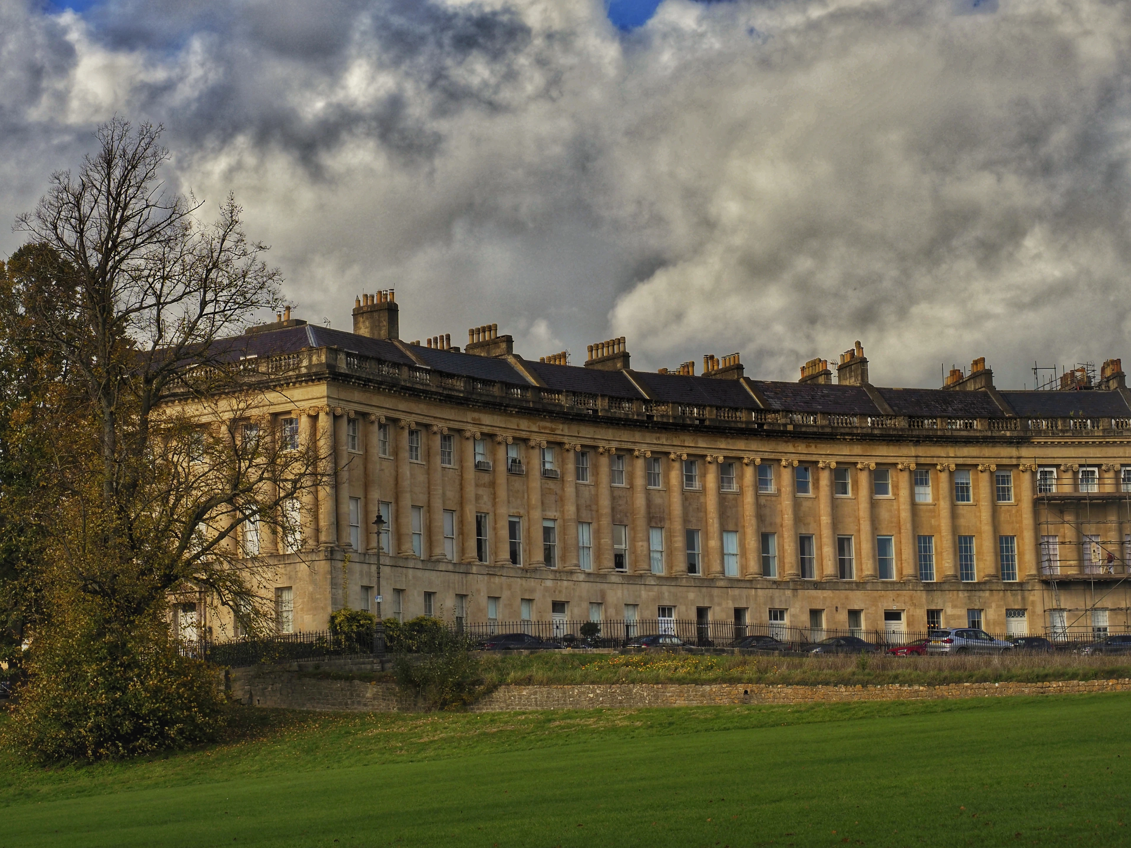 The sweeping Georgian facade of the Royal Crescent in Bath, fronted by a wide green lawn under a dramatic cloudy sky.