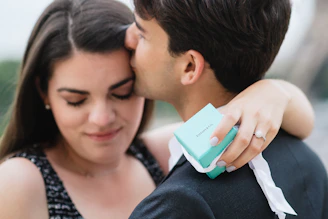A couple shares an intimate moment as a man kisses a woman on the forehead. She is holding a small blue Tiffany box, indicative of an engagement or special occasion, and wears a diamond ring. Her eyes are closed, suggesting contentment, while he appears affectionate.