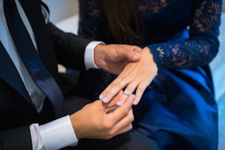 A person wearing a suit places a ring on another person's finger, who is dressed in a blue, lace-patterned outfit. The setting suggests a formal or significant occasion.