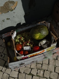 A box filled with various fruits, including a large green melon, red and green grapes, red tomatoes, and other fruits, rests on a cobblestone surface. The carton appears to be slightly worn, and a crumpled paper or plastic bag is also visible inside. The background includes a wall with peeling paint.
