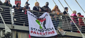 A group of people standing on a bridge with a banner displaying the Palestinian flag and the words 'Stand with Palestine'. Several individuals are holding or photographing the scene, with one person holding a small flag. The crowd appears diverse with people dressed in various styles.