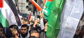 A group of people gathered in a protest. Several flags, including a prominent one with red, white, green, and black colors, are being held by the crowd. The atmosphere appears crowded, with individuals holding up phones to capture the event. Urban surroundings with buildings and traffic lights are visible in the background.