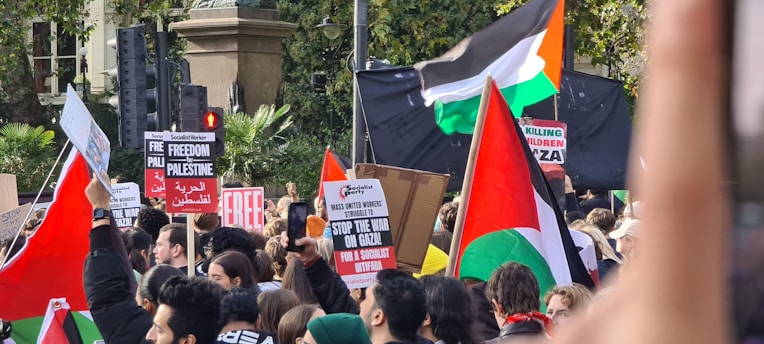 A large group of people are gathered in a protest. They are holding multiple signs and banners with messages such as 'Free Palestine,' 'Stop the War on Gaza,' and 'Stop Killing Children in Gaza.' Several flags can be seen, including the flag of Palestine. The crowd appears to be very engaged and focused on the protest.