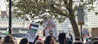 A crowd of people gather holding placards with messages related to Palestinian solidarity. The background features a tree and several buildings. Banners include text such as 'Free Palestine' and 'End Israeli Apartheid.' The scene appears to be a public demonstration.