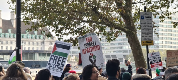 A crowd of people gather holding placards with messages related to Palestinian solidarity. The background features a tree and several buildings. Banners include text such as 'Free Palestine' and 'End Israeli Apartheid.' The scene appears to be a public demonstration.