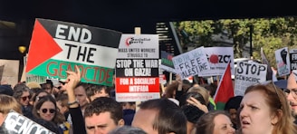 A large group of people are gathered in a protest, holding various signs with messages such as 'End the Genocide,' 'Stop the War on Gaza,' and 'Free Palestine.' The protest appears to be taking place outdoors, with trees visible in the background. Many of the signs are colorful and contain strong political statements. Some protesters are wearing sunglasses and casual clothing.