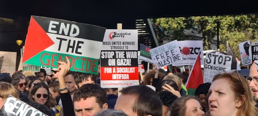 A large group of people are gathered in a protest, holding various signs with messages such as 'End the Genocide,' 'Stop the War on Gaza,' and 'Free Palestine.' The protest appears to be taking place outdoors, with trees visible in the background. Many of the signs are colorful and contain strong political statements. Some protesters are wearing sunglasses and casual clothing.