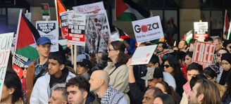 A large group of people gathered in a public space for a protest. Many individuals are holding up signs with messages such as 'Stop the war on Gaza', 'Free Palestine', 'Stop the massacre', and 'War Criminals'. The crowd appears to be diverse, with men and women of different ages. Some people are waving flags and wearing headscarves.