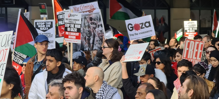 A large group of people gathered in a public space for a protest. Many individuals are holding up signs with messages such as 'Stop the war on Gaza', 'Free Palestine', 'Stop the massacre', and 'War Criminals'. The crowd appears to be diverse, with men and women of different ages. Some people are waving flags and wearing headscarves.