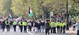 A street is filled with a crowd of people, many of whom are gathered for a demonstration. Several individuals are holding flags, while others are speaking to or observing the gathering. Police officers, identifiable by their bright yellow vests, are present, ensuring the event is orderly. Trees with green leaves line the street, suggesting an urban park area.