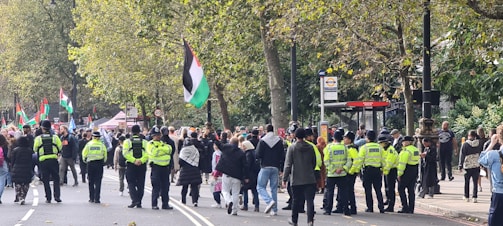 A street is filled with a crowd of people, many of whom are gathered for a demonstration. Several individuals are holding flags, while others are speaking to or observing the gathering. Police officers, identifiable by their bright yellow vests, are present, ensuring the event is orderly. Trees with green leaves line the street, suggesting an urban park area.