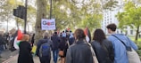 A group of people are participating in a protest or demonstration, carrying signs and flags. Trees and a large building are visible in the background. The atmosphere appears to be lively and engaged.