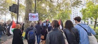 A group of people are participating in a protest or demonstration, carrying signs and flags. Trees and a large building are visible in the background. The atmosphere appears to be lively and engaged.