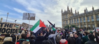 A large group of people gathered in a protest outside an ornate building, holding flags and signs with messages of support for Palestine. The crowd appears diverse, with many individuals facing the building, contributing to a sense of unity and determination.