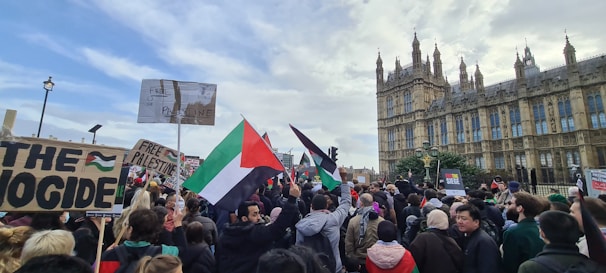A large group of people gathered in a protest outside an ornate building, holding flags and signs with messages of support for Palestine. The crowd appears diverse, with many individuals facing the building, contributing to a sense of unity and determination.