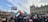 A large group of people gathered in a protest outside an ornate building, holding flags and signs with messages of support for Palestine. The crowd appears diverse, with many individuals facing the building, contributing to a sense of unity and determination.