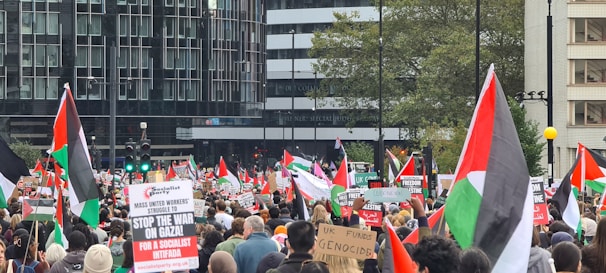 A large crowd is gathered for a protest, holding flags and signs with slogans related to social and political issues. The setting is urban, with tall buildings and trees in the background.