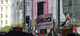 A protest scene with people holding various signs and flags. The central sign reads 'Gaza Stop the massacre.' Other signs and slogans are partially visible. A variety of flags are present, and the setting is urban with large glass buildings and a traffic light in the background.