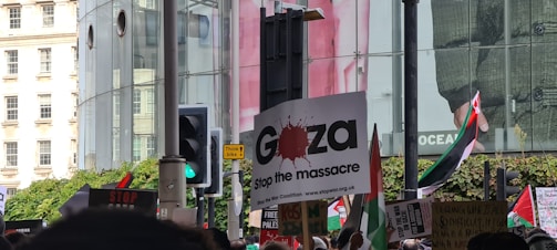 A protest scene with people holding various signs and flags. The central sign reads 'Gaza Stop the massacre.' Other signs and slogans are partially visible. A variety of flags are present, and the setting is urban with large glass buildings and a traffic light in the background.