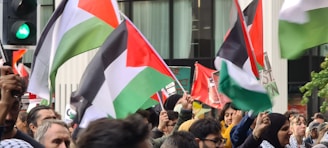 A crowd of individuals is gathered, holding Palestinian flags. The flags display horizontal stripes of black, white, and green with a red triangle. People are waving the flags energetically, and there is a traffic light visible, displaying a green signal. Some banners and slogans are partially obscured by the crowd.