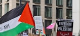 A protest scene featuring multiple signs and flags, including one with the colors of the Palestinian flag. Signs contain messages related to the Palestinian freedom movement. The background includes a modern building façade.