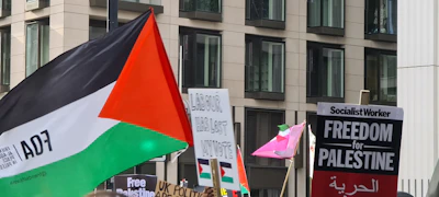 A close-up of a protest sign with a Palestinian-style flag waving in the wind near a city square.