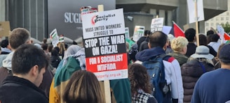 A crowd of people participating in a protest, holding various signs that express opposition to the war in Gaza. The central sign calls for a socialist intifada, organized by the Socialist Party. Many participants are wearing jackets and backpacks.