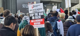 A crowd of people participating in a protest, holding various signs that express opposition to the war in Gaza. The central sign calls for a socialist intifada, organized by the Socialist Party. Many participants are wearing jackets and backpacks.