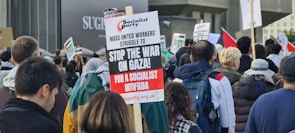 A crowd of people participating in a protest, holding various signs that express opposition to the war in Gaza. The central sign calls for a socialist intifada, organized by the Socialist Party. Many participants are wearing jackets and backpacks.