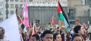 A large group of people gather in front of a building with banners and flags, including red, green, and black flags. Some participants hold signs with messages supporting Palestine. The scene appears to be a demonstration or protest.