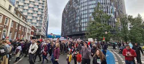 A large group of people, including adults and children, are participating in a street protest or march. Many individuals are holding flags and signs with various messages. The crowd is walking along a city street flanked by modern and older buildings. Trees line part of the street, and the mood appears engaged and solemn.