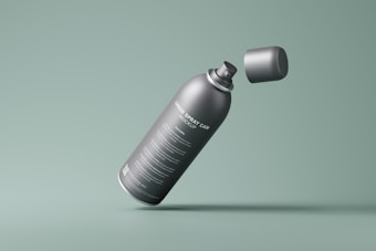 A grey spray can with detailed text labeling on it is prominently displayed against a muted green background. The cap of the spray can appears to be floating in mid-air, separated from the body of the can.