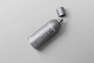 A gray spray can with a detachable cap lying on its side on a light gray surface. The can has text written on it in white color.