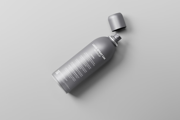 A gray spray can with a detachable cap lying on its side on a light gray surface. The can has text written on it in white color.