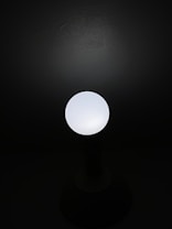 A single glowing light bulb emits a soft white light against a dark background, creating a stark contrast between light and shadow.