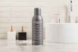 A gray spray can prominently placed on a polished bathroom countertop. The can has text indicating it is a mockup, with several features listed beneath. In the background, a white circular sink with a chrome faucet is visible, alongside two soap dispensers, one white and one black. The wall is tiled with light beige bricks, giving a clean and modern look.