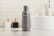 A gray spray can prominently placed on a polished bathroom countertop. The can has text indicating it is a mockup, with several features listed beneath. In the background, a white circular sink with a chrome faucet is visible, alongside two soap dispensers, one white and one black. The wall is tiled with light beige bricks, giving a clean and modern look.