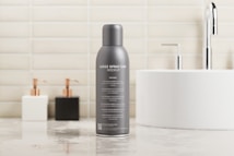 A gray spray can prominently placed on a polished bathroom countertop. The can has text indicating it is a mockup, with several features listed beneath. In the background, a white circular sink with a chrome faucet is visible, alongside two soap dispensers, one white and one black. The wall is tiled with light beige bricks, giving a clean and modern look.