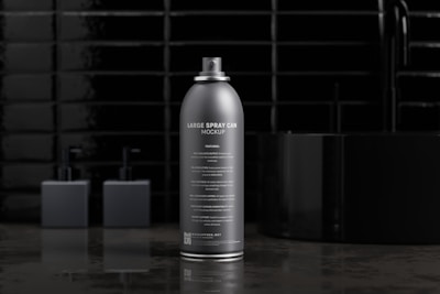 A sleek, silver spray can is prominently displayed on a reflective surface. The background features a black glossy tiled wall and blurred shapes that resemble containers, adding to the minimalist aesthetic.