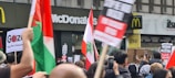 Crowd of people holding various protest signs and flags in front of McDonald's and Itsu restaurants. Some signs have messages related to stopping a massacre and proclaiming freedom for Palestine.